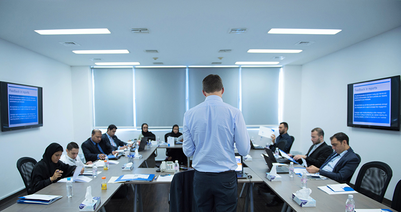 A number of people sit at a row of desks while a meeting takes place